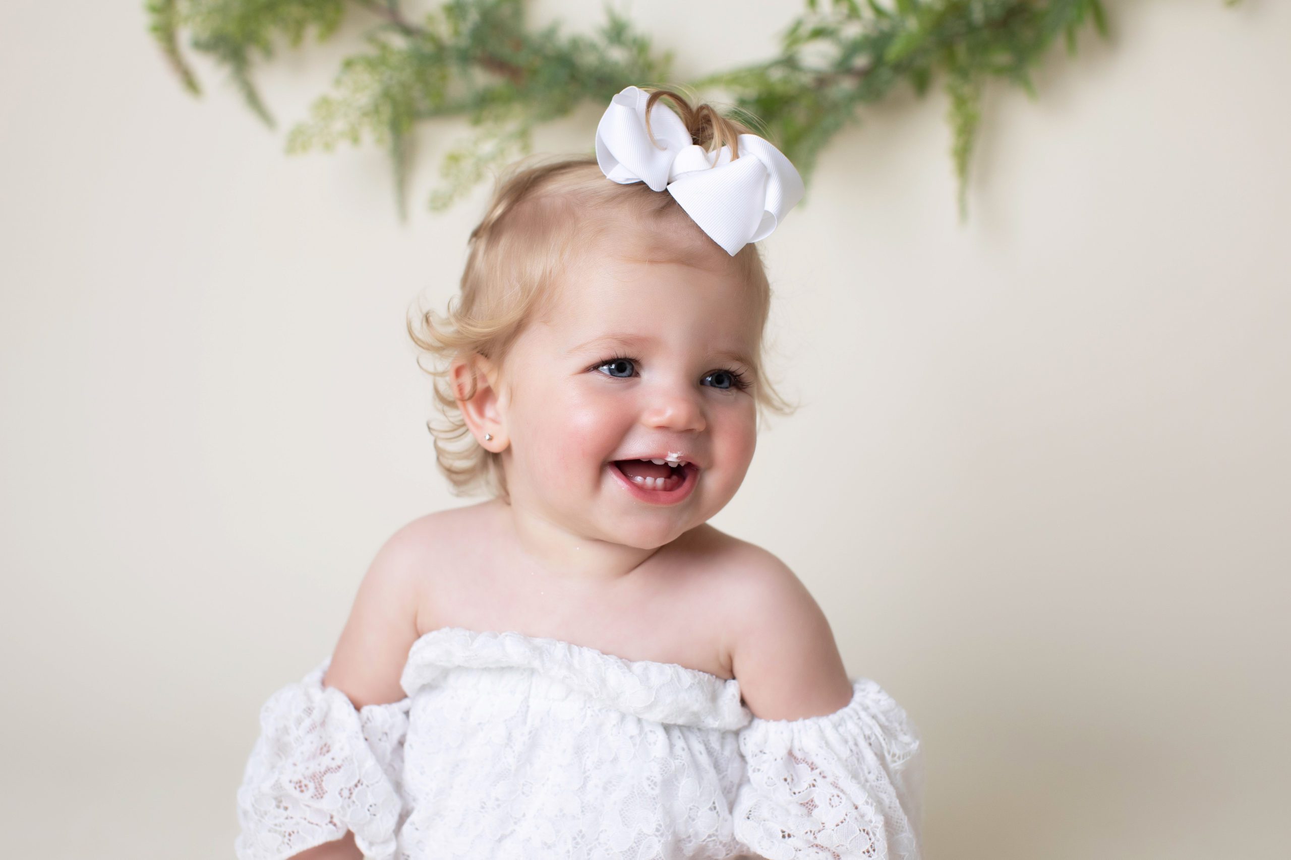 Smiling baby girl with blue eyes and white bow in a lace off-shoulder dress during a Fairfax, VA milestone studio session.