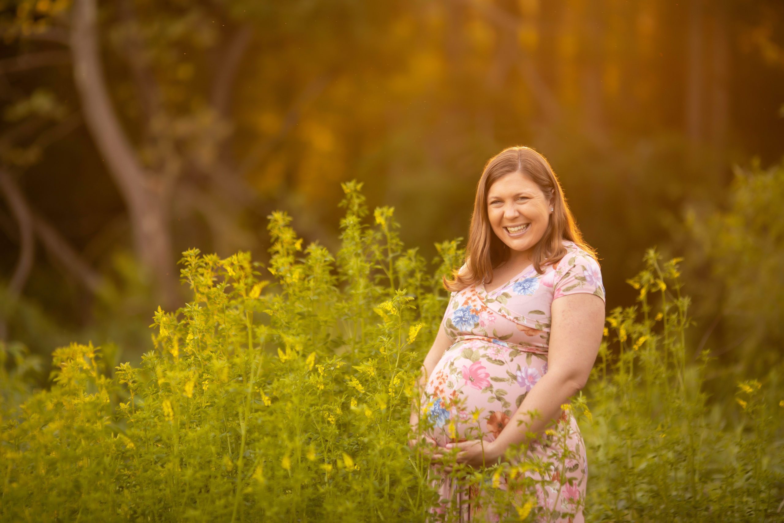 Pregnant woman holding her baby bump while standing in tall yellow wildflowers, bathed in warm sunset light in Fairfax, VA.