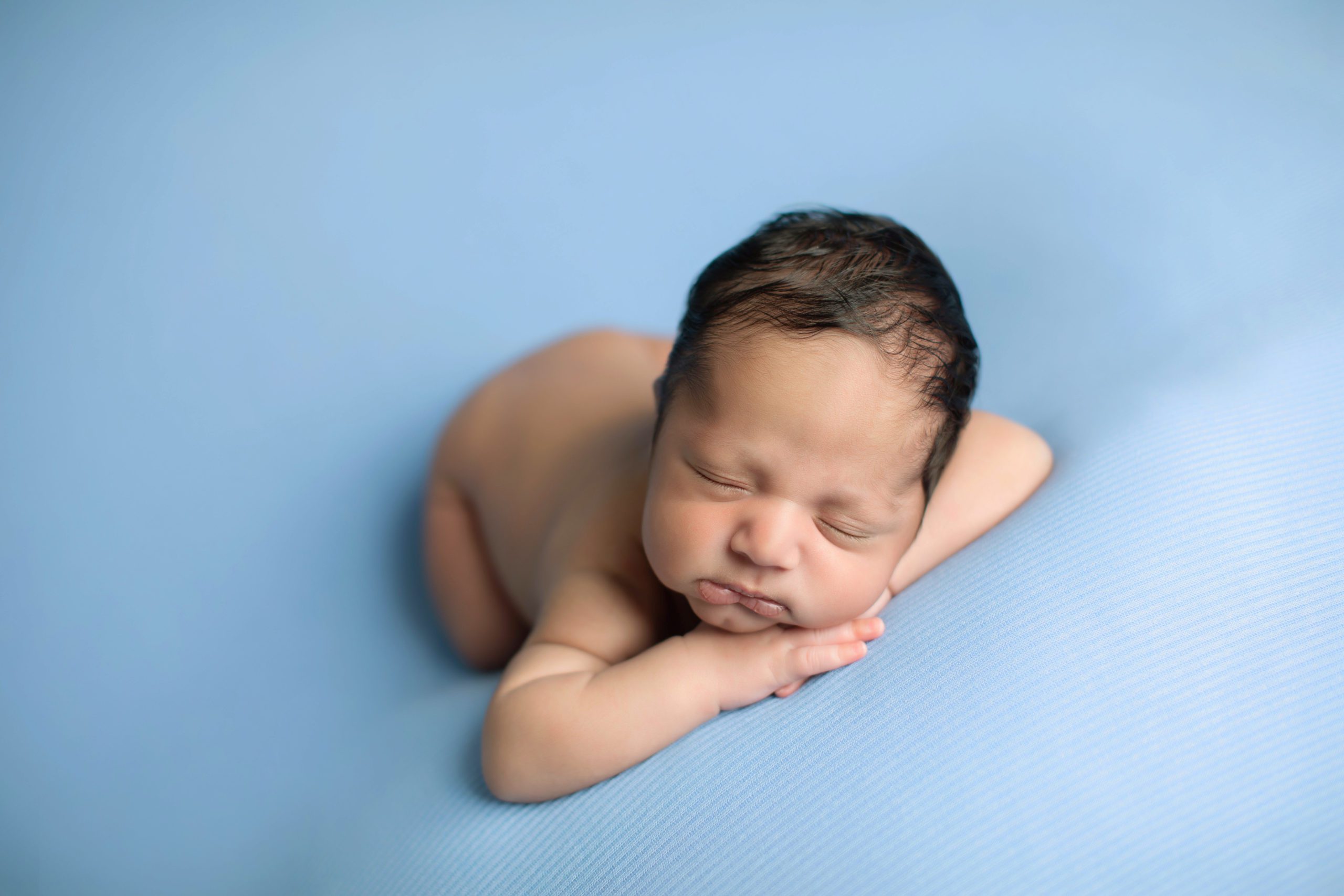 Newborn baby boy lying peacefully on a blue blanket with natural light highlighting his delicate features. Photographed by Marian Lozano in Fairfax, VA.